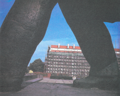 A building under construction seen in the distance through the legs of a statue
