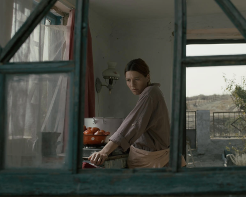 A still from the film "Klondike" of a woman in her kitchen as seen through a window